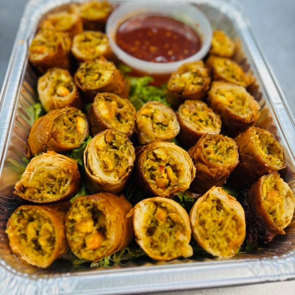 A catering tray of golden-brown Mediterranean spring rolls filled with vegetables and chicken, with a side of dipping sauce.
