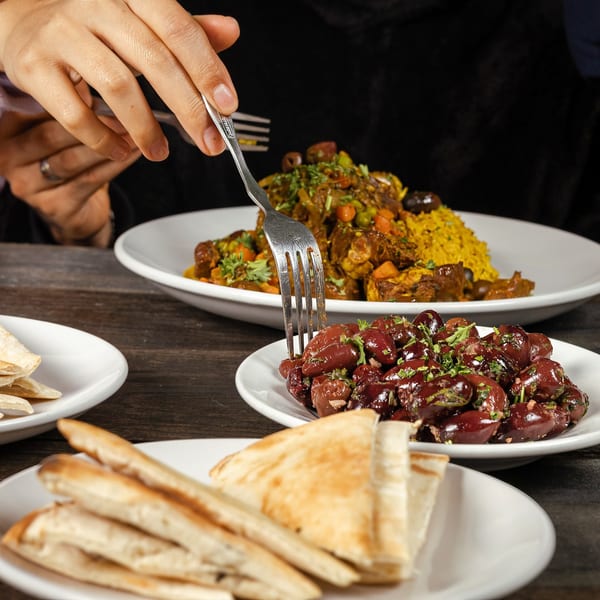 A person's hand forks a Mediterranean olive appetizer, with a main dish of meat and rice in the background.