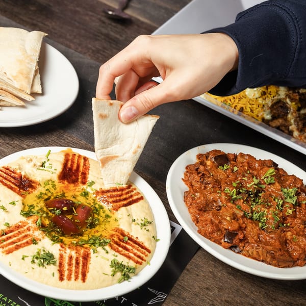 A hand dips pita bread into a bowl of hummus, with another Mediterranean dish beside it.