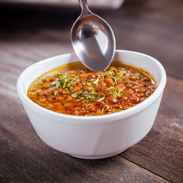 A spoon dips into a white bowl of Mediterranean lentil soup garnished with fresh herbs.